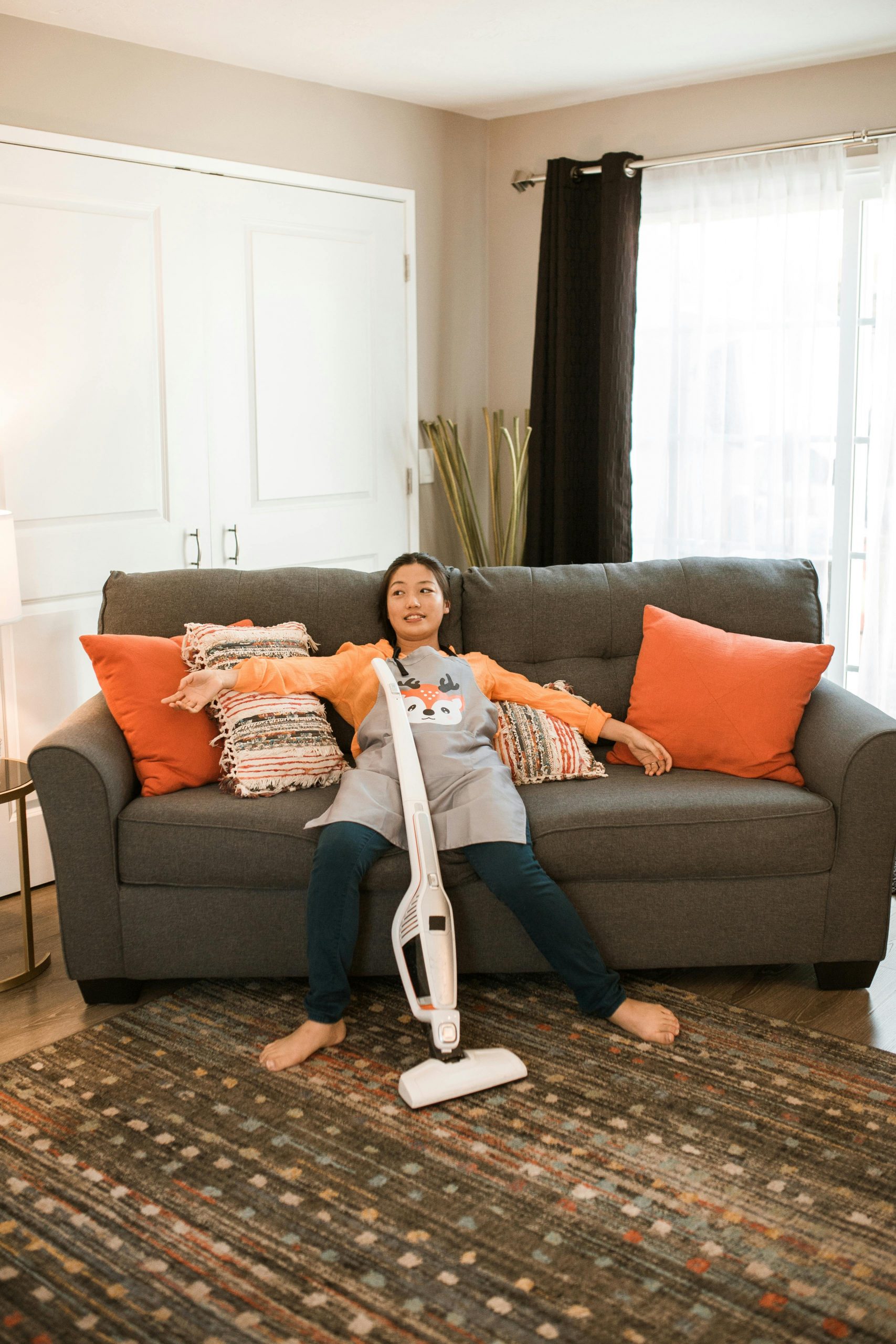 Asian woman resting on a couch indoors with a vacuum cleaner after cleaning.